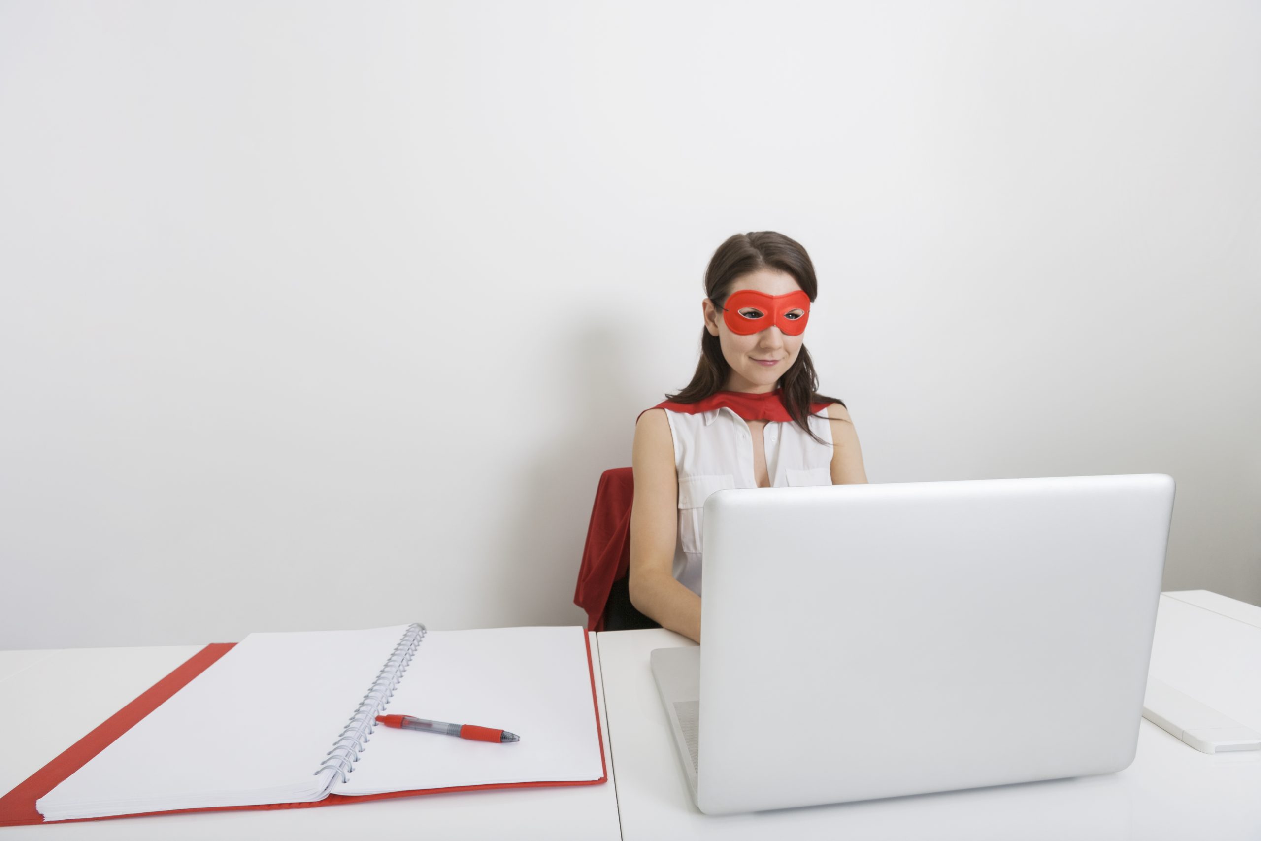 Young businesswoman dressed as superhero using laptop at desk in office