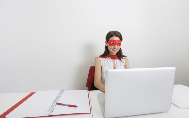 Young businesswoman dressed as superhero using laptop at desk in office